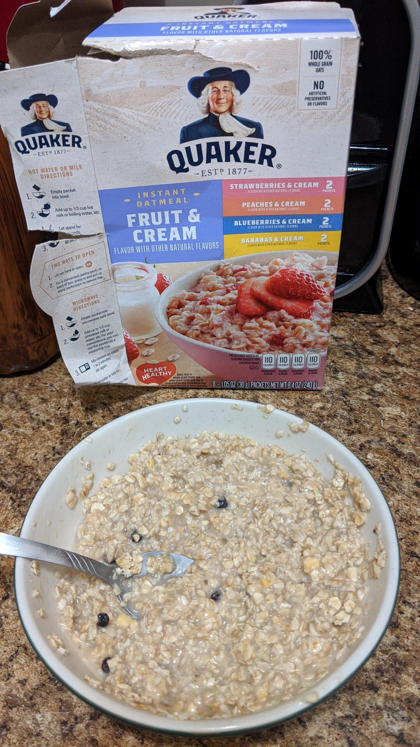 A bowl of oatmeal with a spoon sits on a counter in front of a Quaker Fruit & Cream oatmeal box. Some blueberries are visible in the oatmeal.