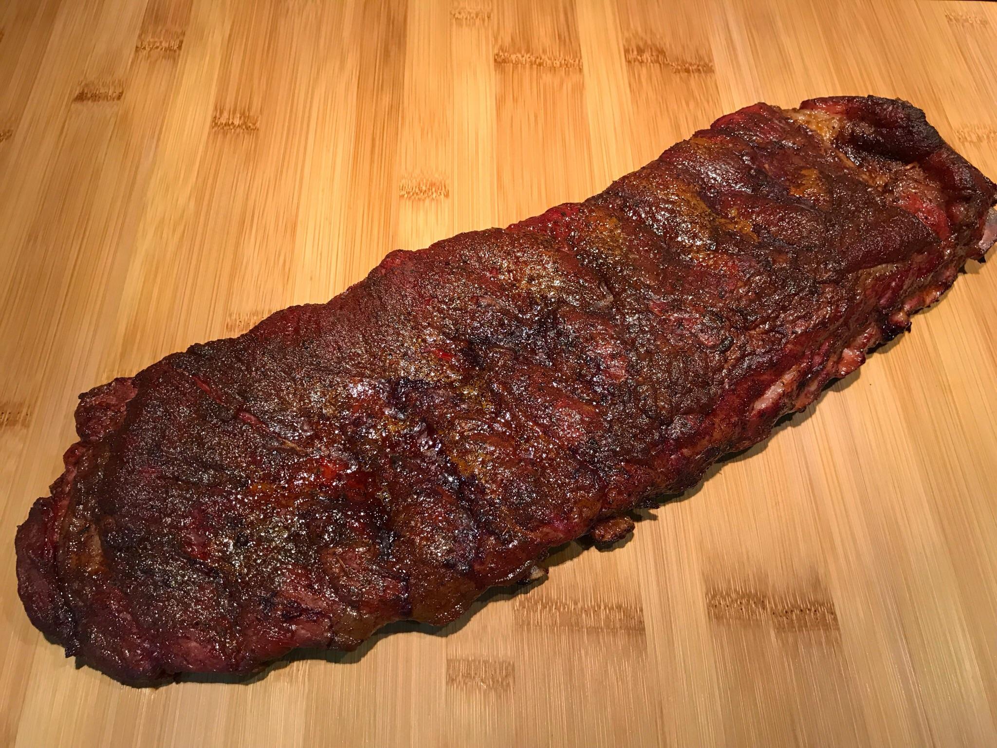 A full slab of cooked, seasoned barbecue ribs rests on a wooden cutting board, displaying a dark, caramelized crust.