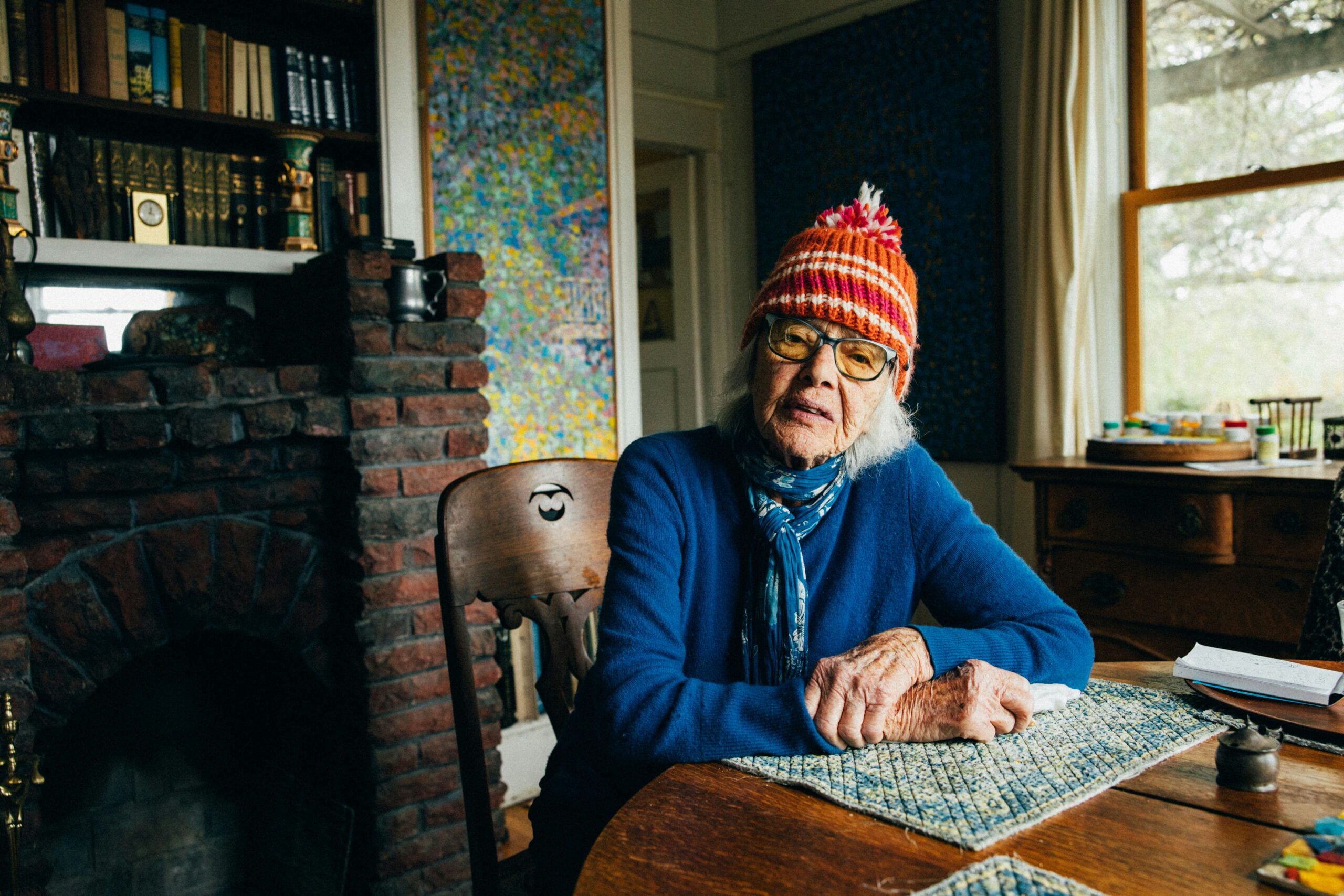 An elderly woman wearing glasses and a colorful knit hat sits at a table in a cozy room with a brick fireplace, bookshelf, and window letting in natural light, perhaps sharing some classic Boomer facts as she enjoys the peaceful atmosphere.