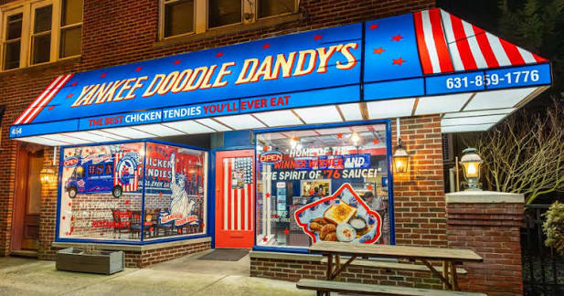 Yankee Doodle Dandy’s restaurant exterior at night, featuring a bright red, white, and blue awning with stars and stripes. Windows display menus and patriotic décor. A wooden bench sits outside.