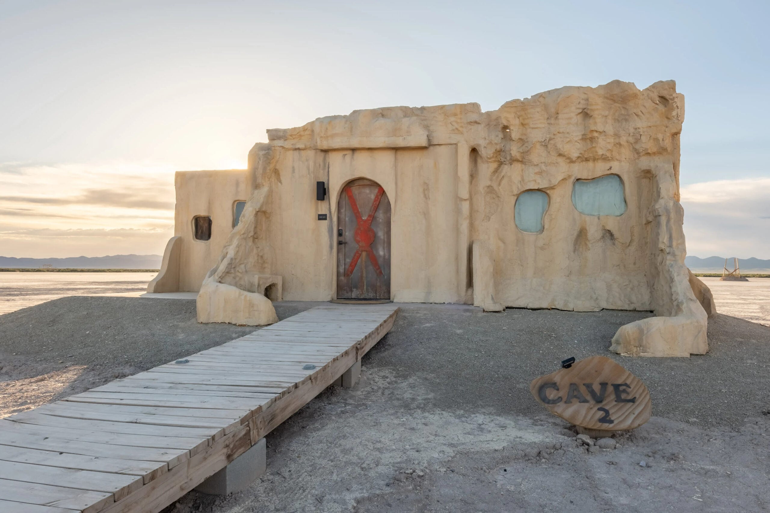 A beige, cave-like building with rounded windows and a red-X marked door stands in a desert. A wooden path leads to the entrance, and a sign reading “CAVE 2” sits on the right. The sun sets in the background.