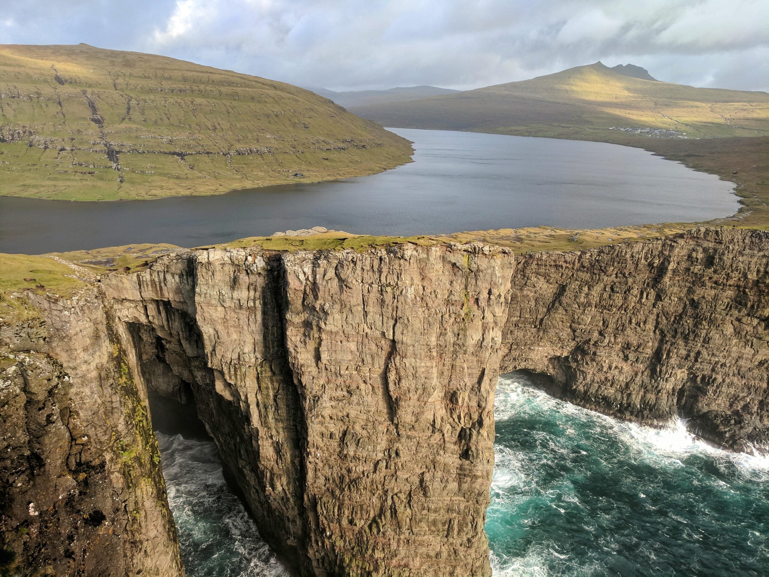 A large cliff rises above the ocean with waves crashing below, and a calm lake sits just behind the cliff, surrounded by grassy hills under a cloudy sky.