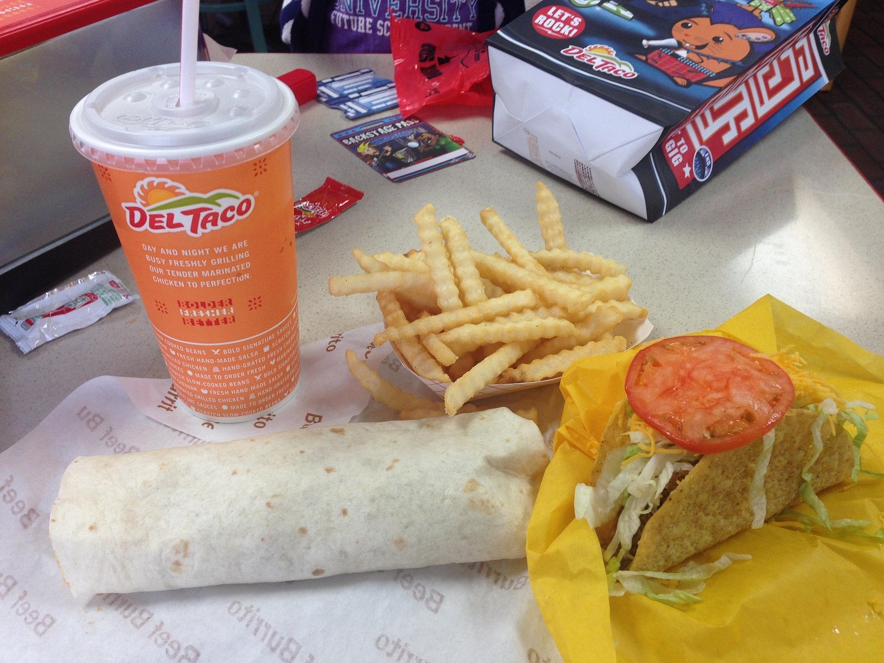 A Del Taco meal with a soda, crinkle-cut fries, a wrapped burrito, and a hard shell taco with lettuce and tomato, all on a table.