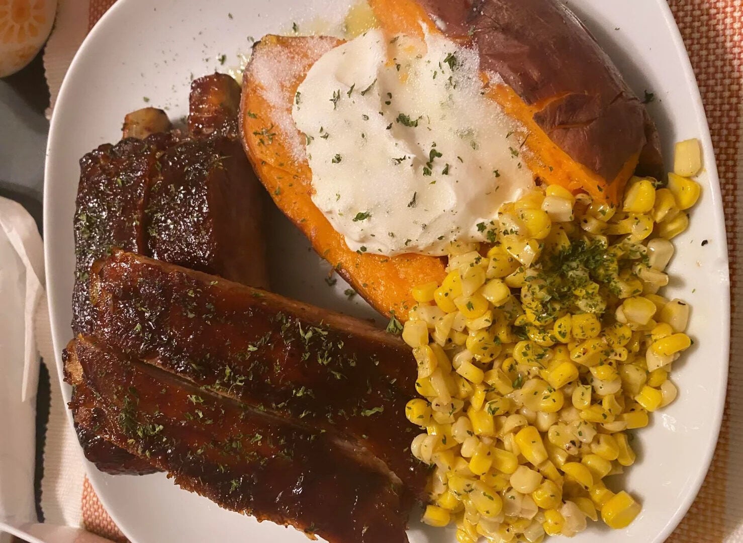 A plate with glazed ribs, a baked sweet potato topped with sour cream and herbs, and a serving of seasoned corn.