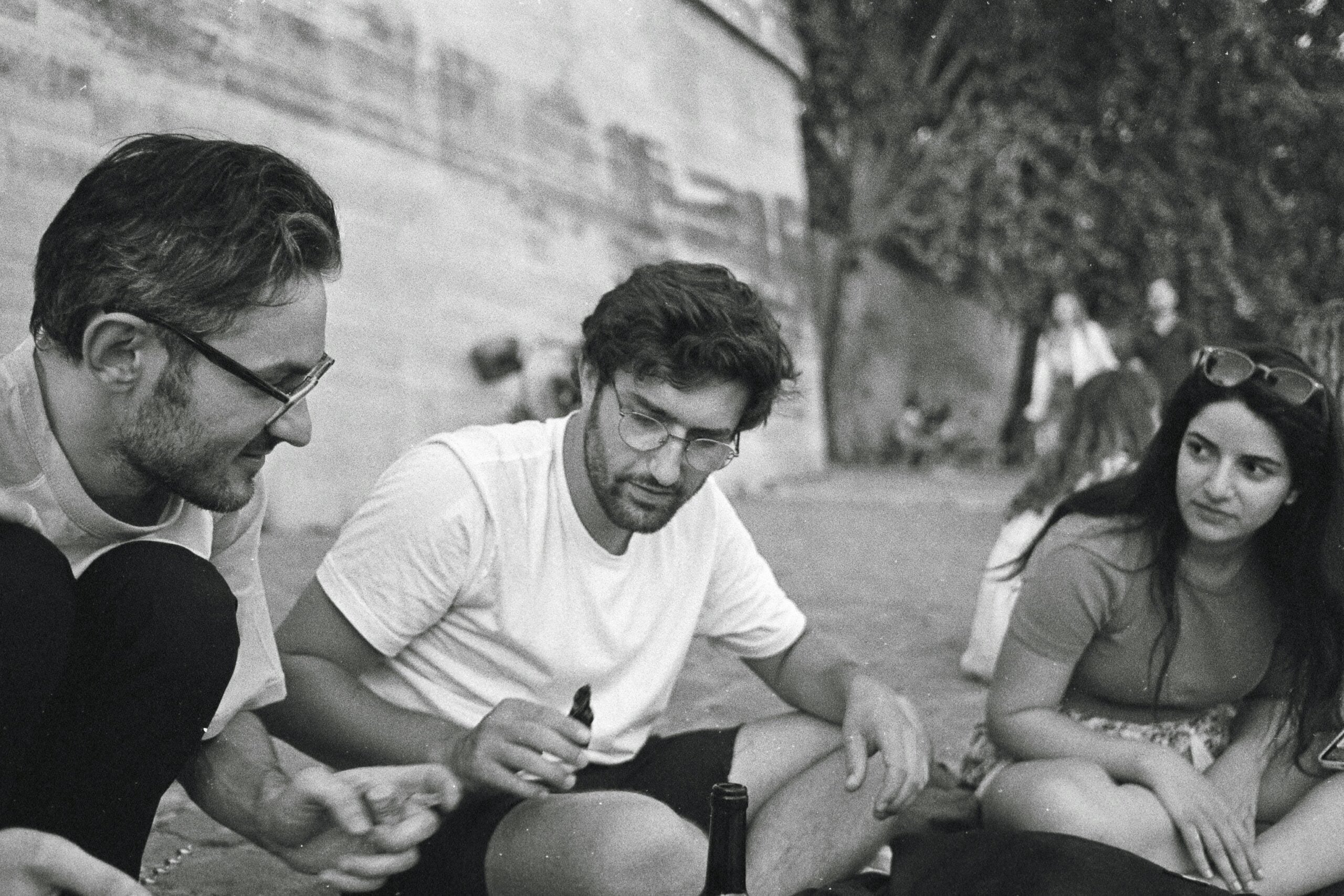 Three people sit outdoors on the ground, engaged in conversation about life in 2026. Two men wear glasses and casual clothes; one woman with long dark hair looks at them. Trees and a stone wall are in the background.
