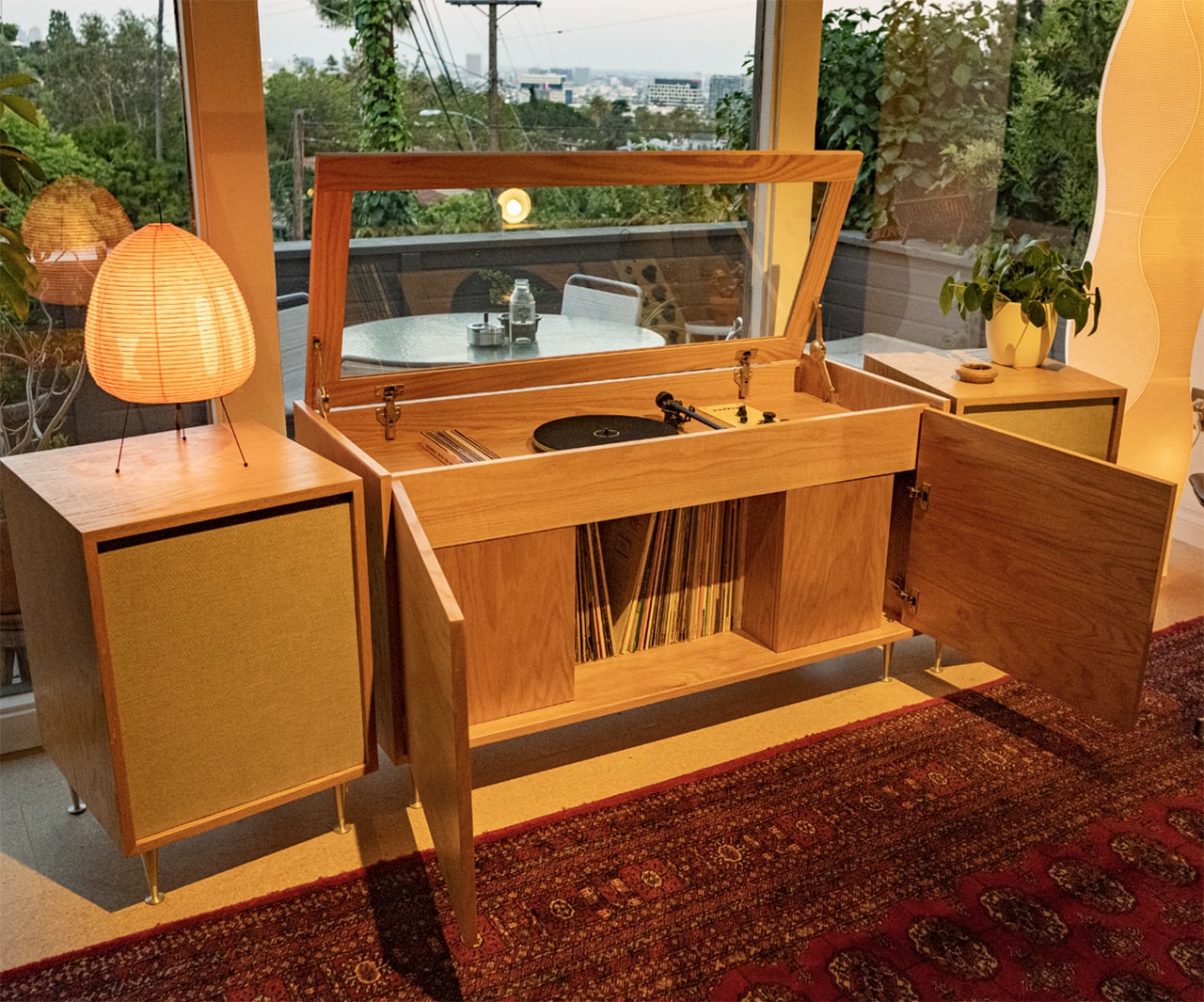A wooden record player console with open doors reveals vinyl records and a turntable. It sits by large windows, next to a lamp, plant, and overlooks an outdoor patio with greenery.