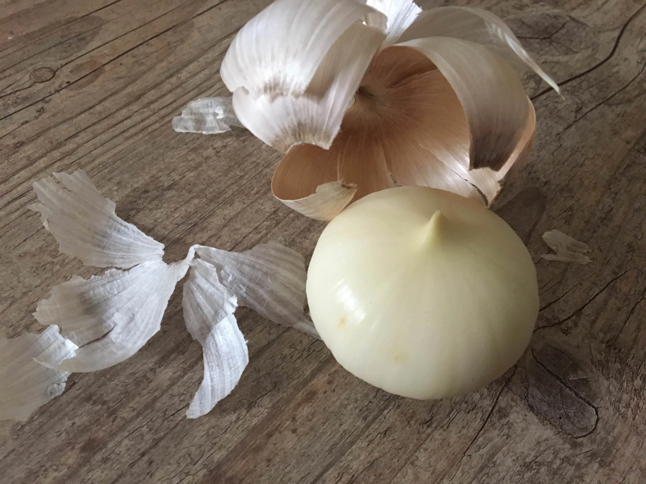 A peeled onion with its papery skin scattered on a wooden surface. The onion sits in the foreground, with some of the skin curled and some pieces flat beside it.