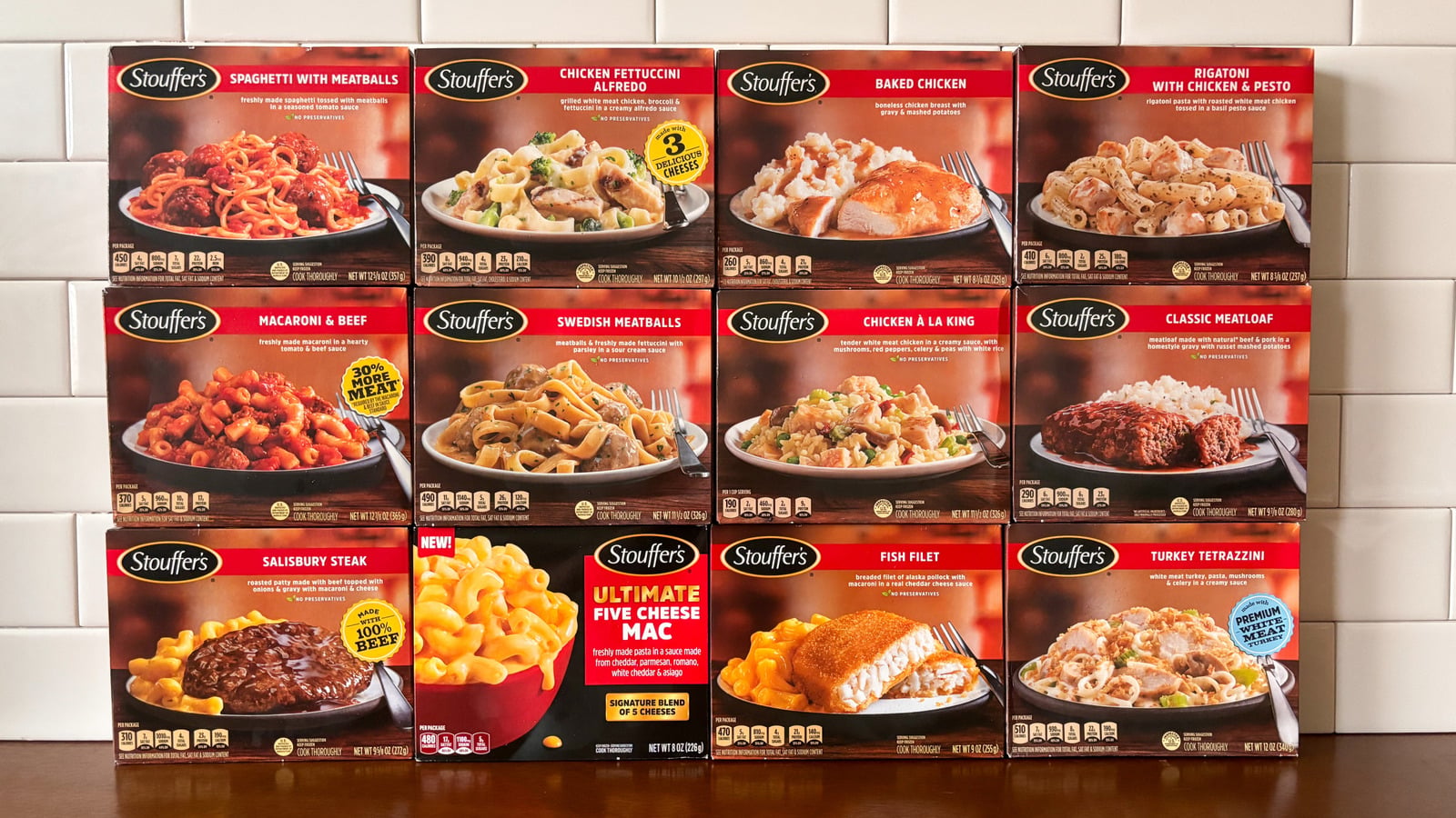 Twelve Stouffer’s frozen meal boxes are arranged in three rows against a white tiled wall, showcasing a variety of entrees like spaghetti, chicken, meatloaf, macaroni, and fish filet.