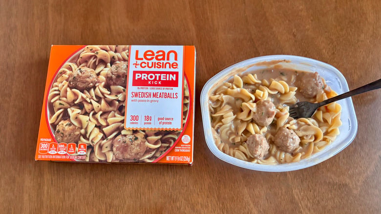 A Lean Cuisine Swedish Meatballs frozen meal box sits next to the prepared meal in a plastic tray with a fork, showing pasta, meatballs, and creamy gravy on a wooden surface.