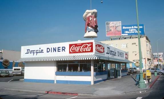 A white diner with blue trim features a large dog head in a chef hat on the roof and a Coca-Cola sign. A billboard and other buildings are visible in the background.