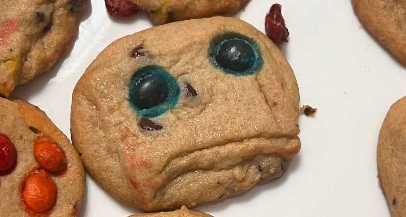 A cookie with two blue candies for eyes and a curved line resembling a sad mouth, giving it a face-like appearance. Other cookies with colored candies are nearby on a white plate.