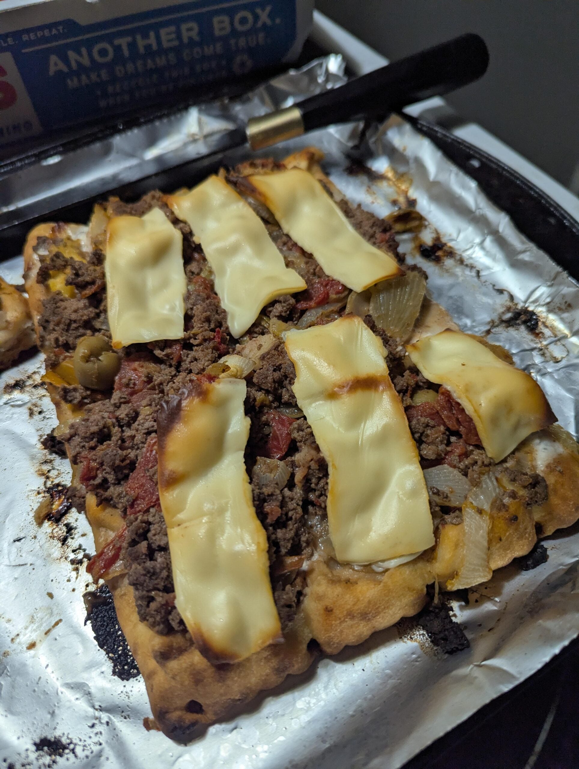 A homemade rectangular pizza on foil, topped with ground beef, onions, olives, and melted cheese slices, with a pizza box visible in the background.