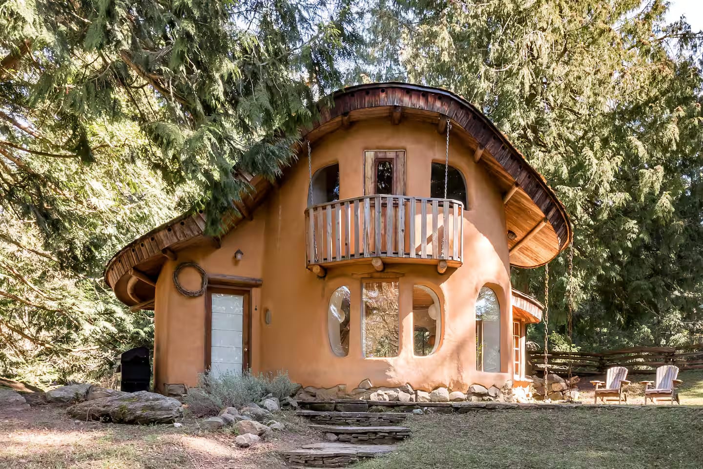 A whimsical, round mud house with curved windows, a small balcony, and a wavy roof, surrounded by trees and outdoor chairs on a grassy area.