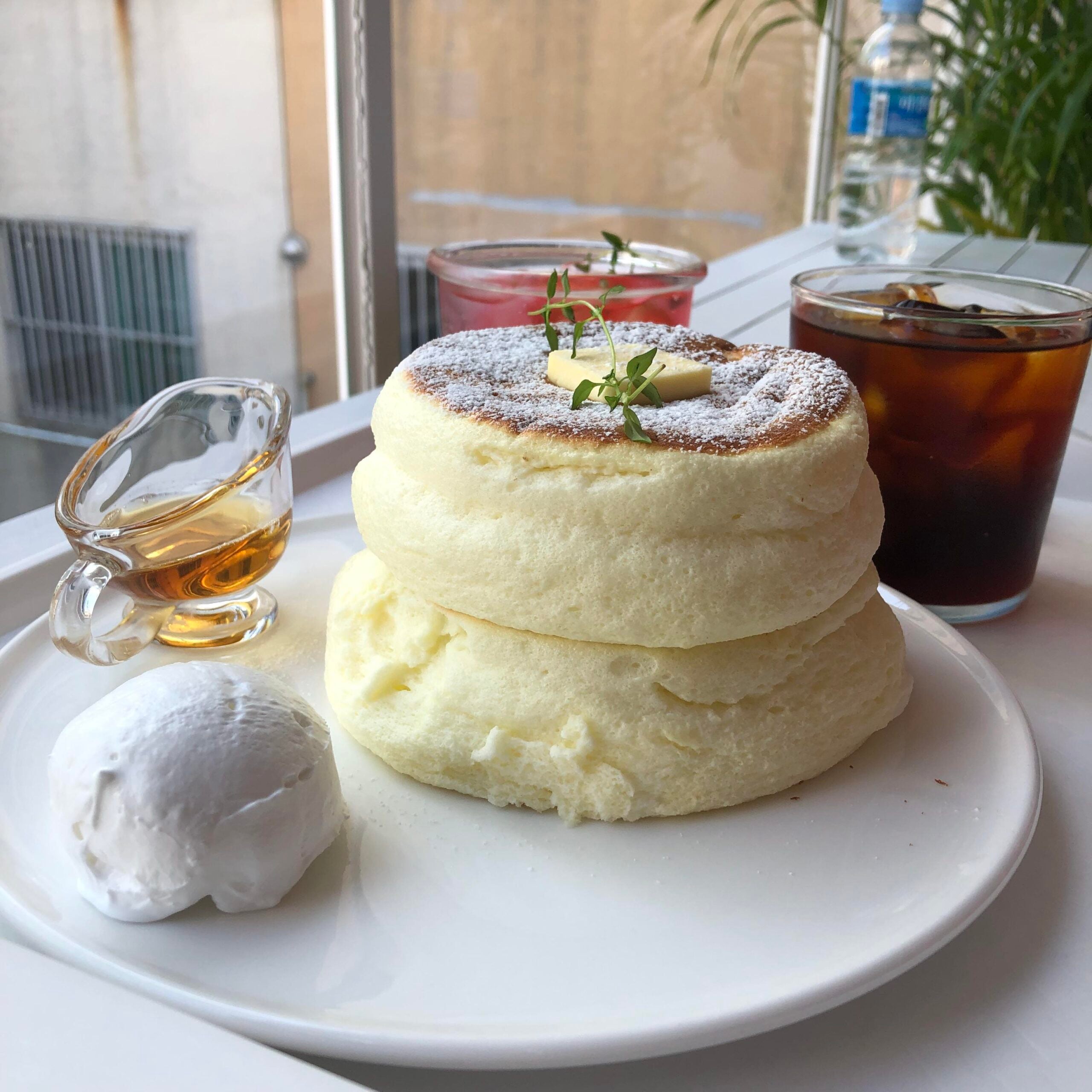 A plate of fluffy souffle pancakes with butter, powdered sugar, syrup, and whipped cream, served with iced coffee and a pink drink by a window.