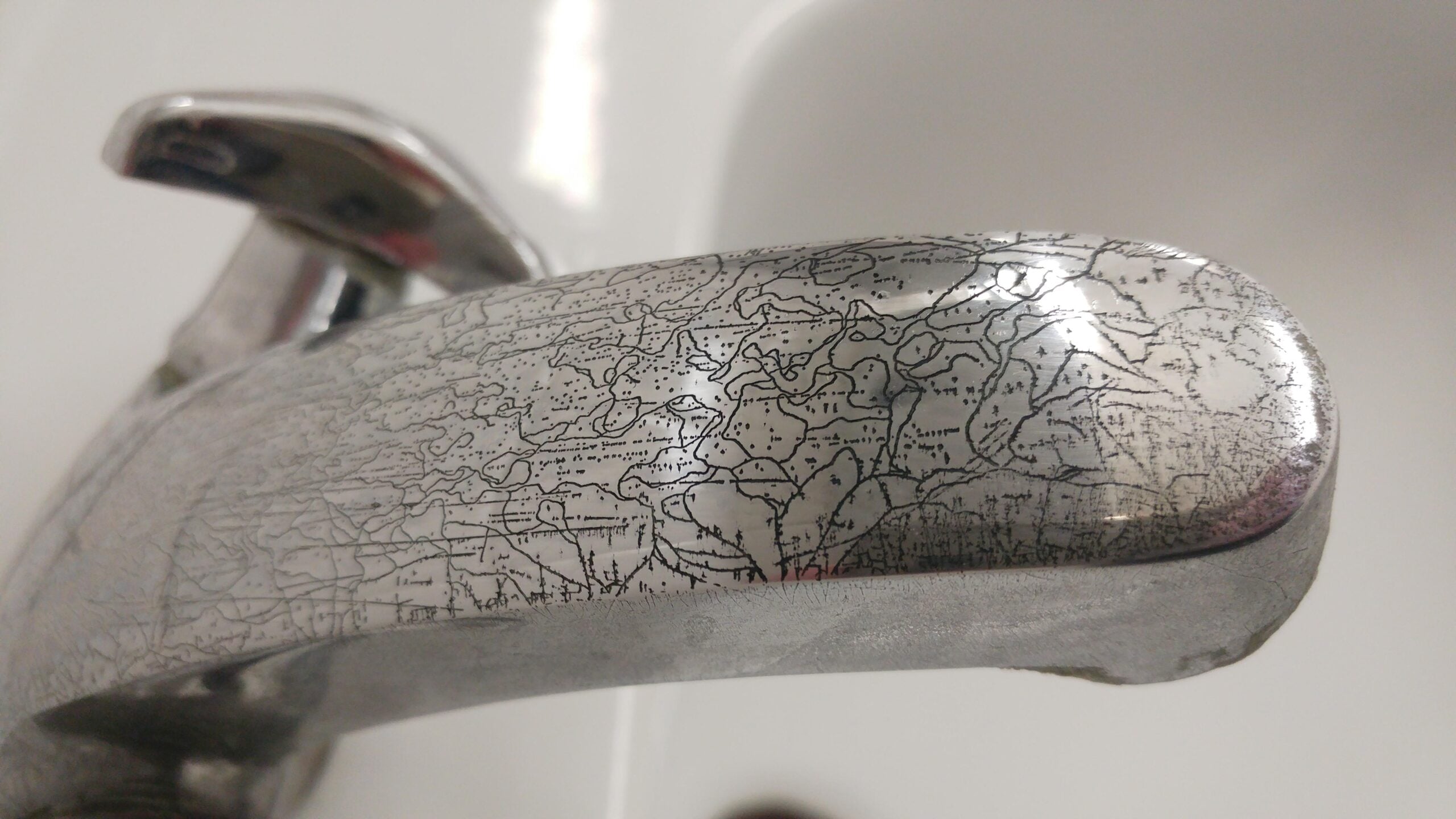 Close-up of a metal faucet handle with a worn, cracked, and tarnished surface, showing intricate patterns and lines. The background is out of focus, featuring a white sink.