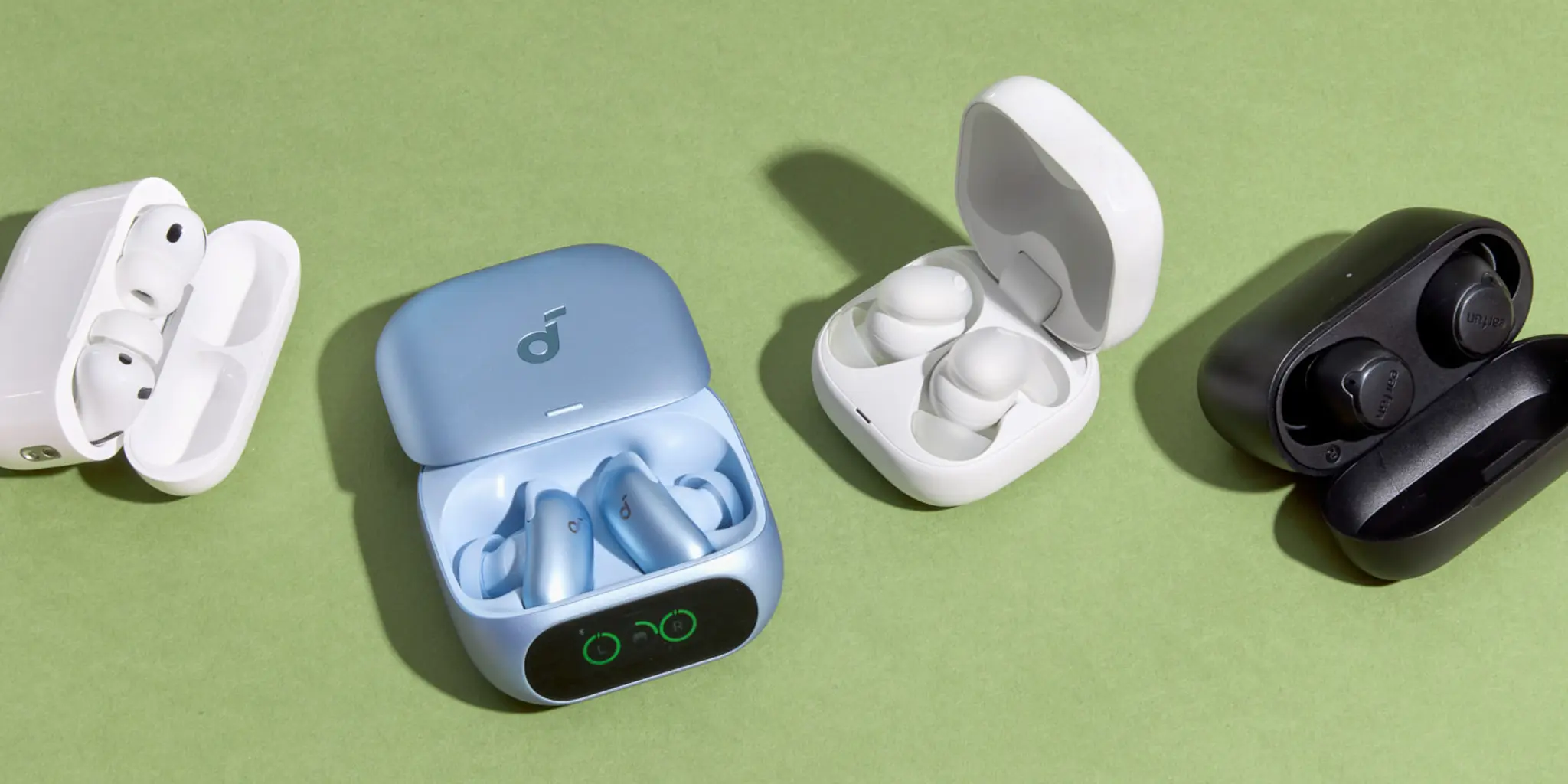 Four pairs of wireless earbuds in different colors and cases—white, blue, white, and black—are displayed on a green surface, with their cases open showing the earbuds inside.