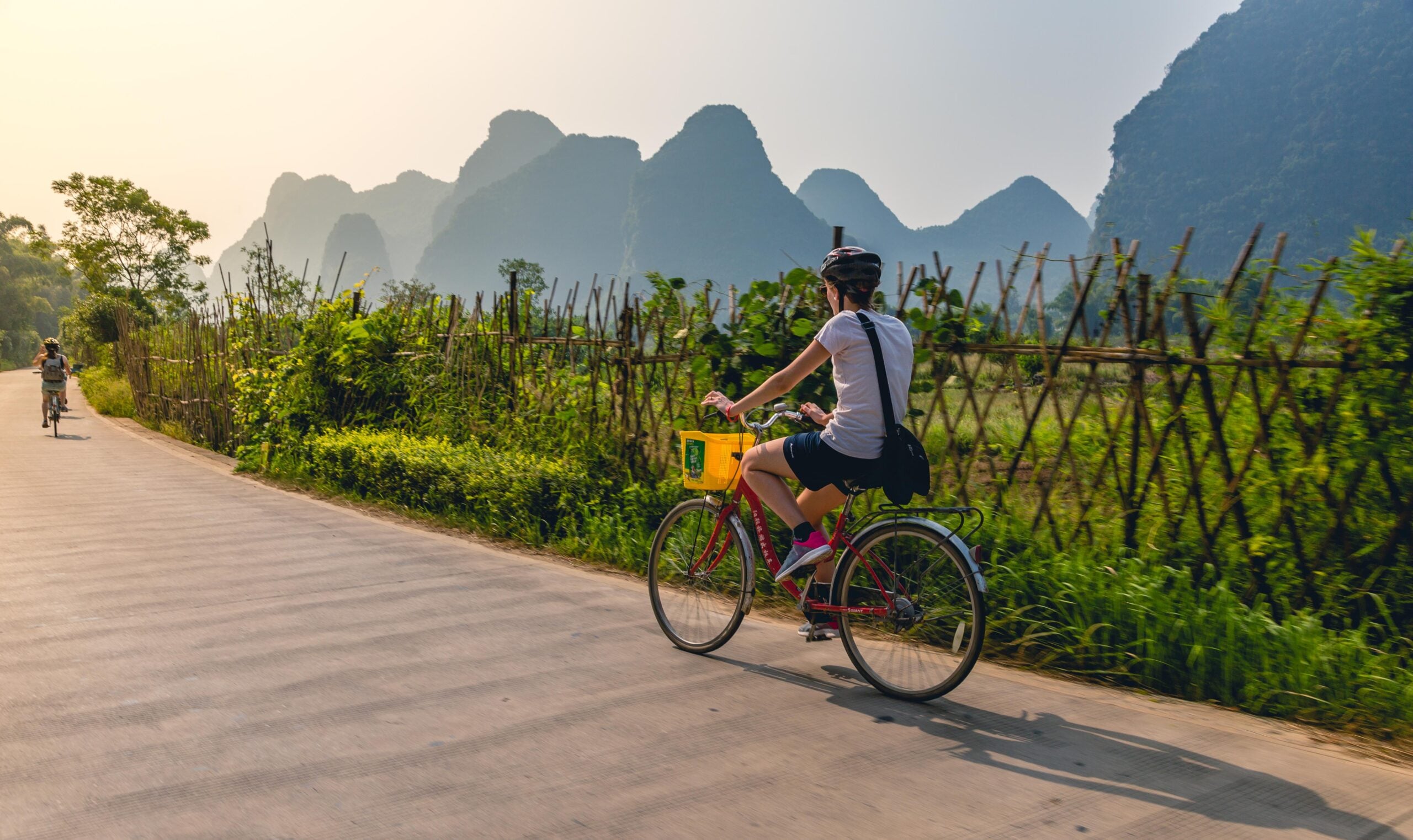 Person riding a bicycle on a paved road next to a bamboo fence, with lush greenery and tall, misty mountains in the background under a clear sky. Another cyclist is visible in the distance.