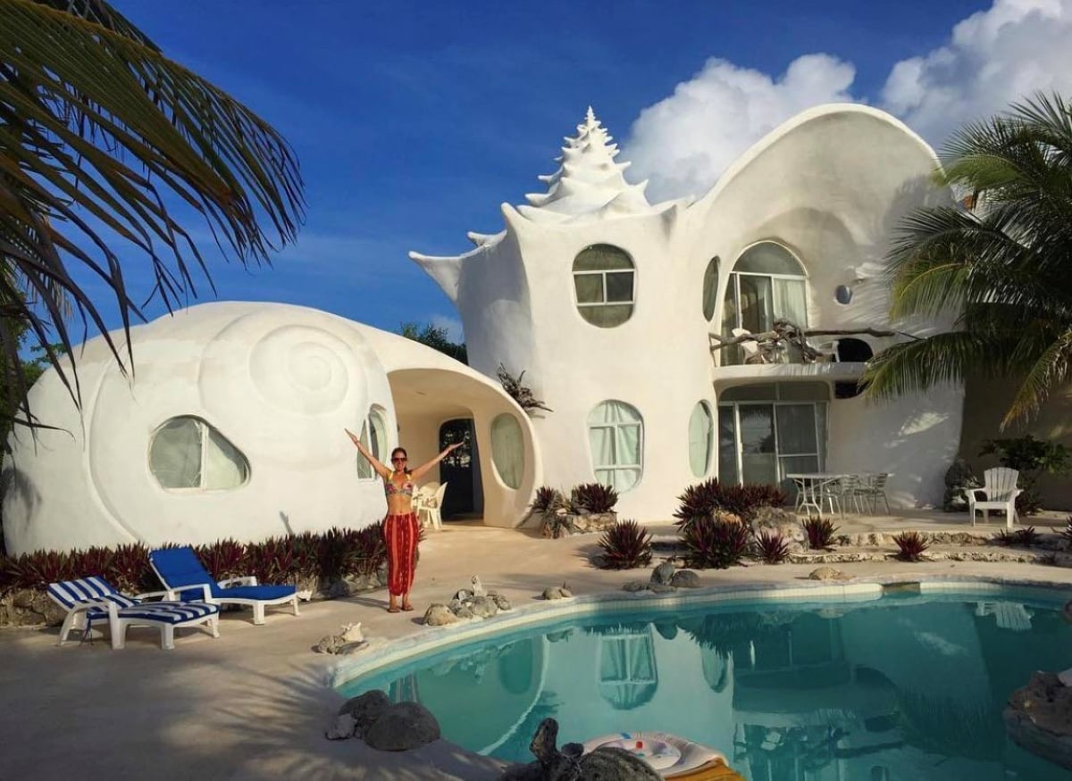 A woman stands by a turquoise pool in front of a unique white house shaped like a seashell, surrounded by tropical plants and palm trees under a bright blue sky.