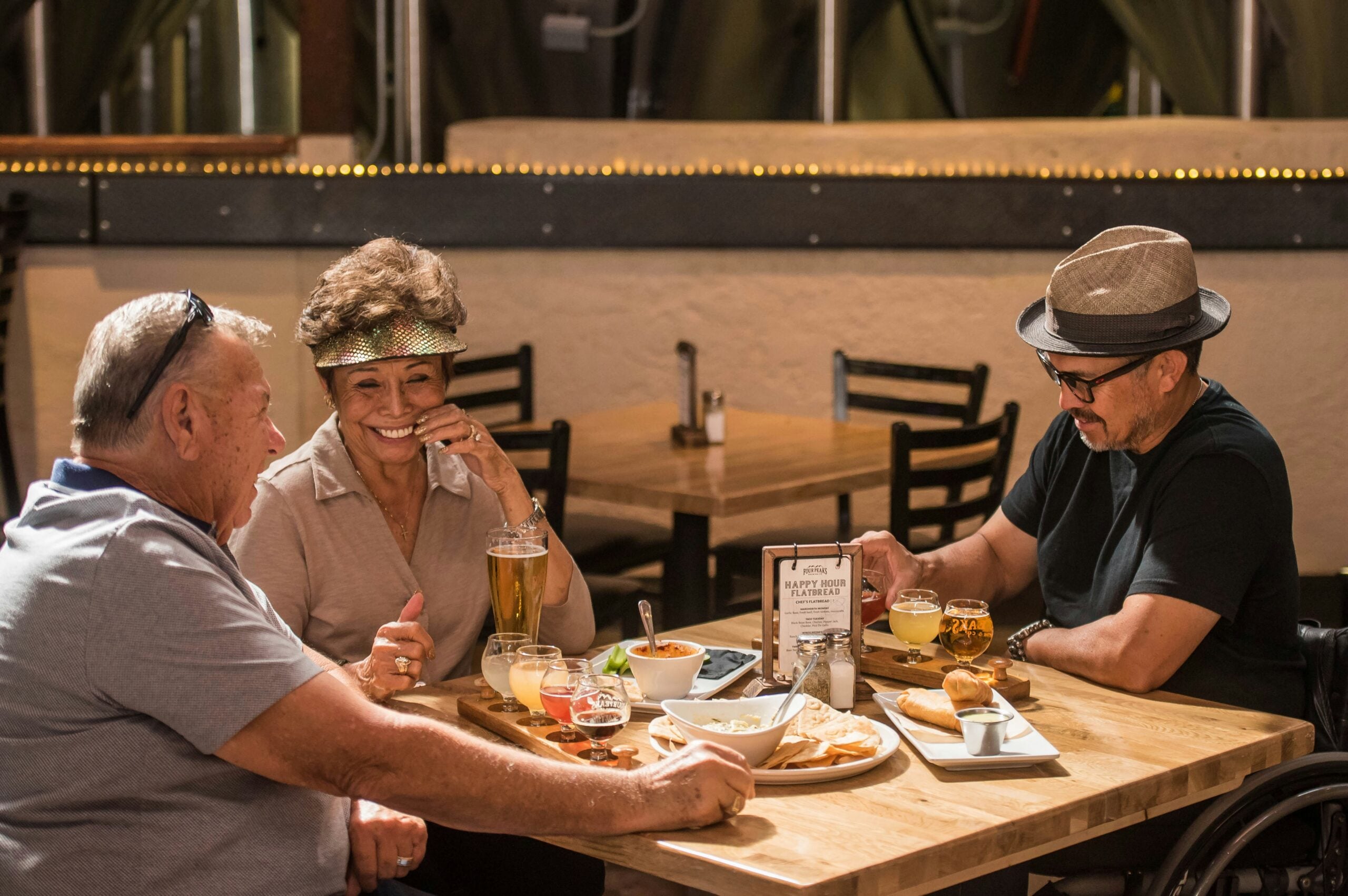 Three older adults sit at a wooden table in a restaurant, laughing and enjoying drinks and food together. Beer flights, snacks, and menus are visible on the table as the group of boomers—clearly experts at saving money—share stories and good times without breaking the bank. The atmosphere is casual and cheerful.
