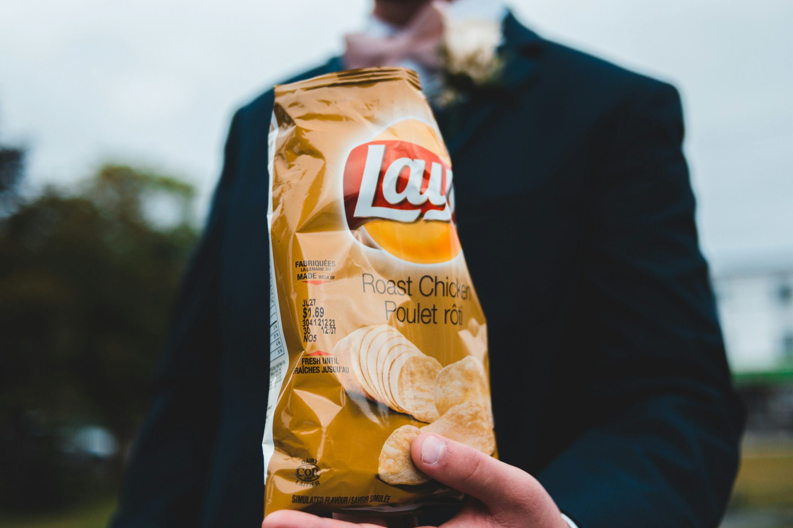 A person in a suit holds a bag of Lay’s Roast Chicken flavored potato chips, with the focus on the chips and the person’s hand, while the background is blurred.