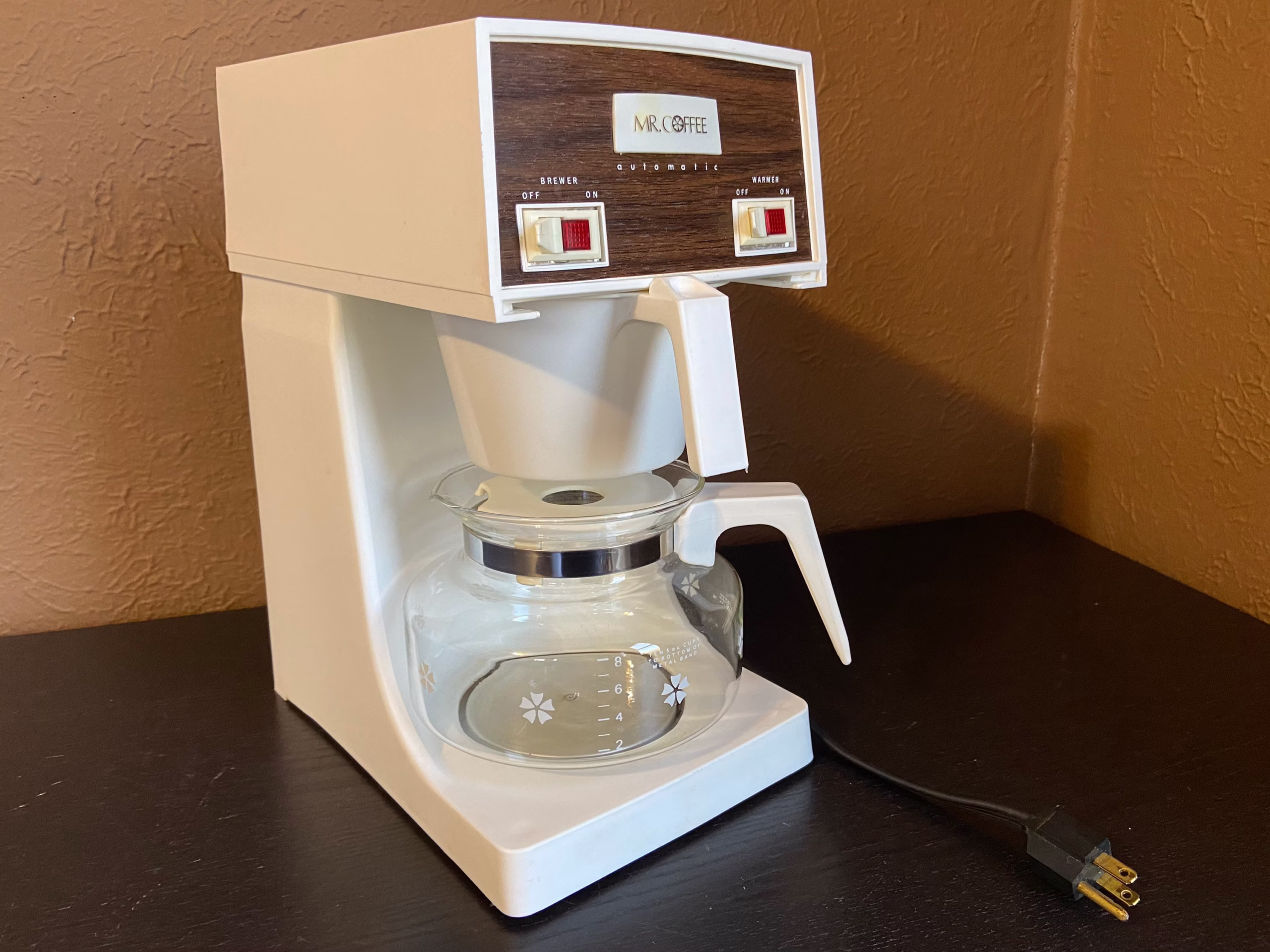 A vintage white Mr. Coffee machine with a glass carafe sits on a dark table against a brown textured wall. The power cord is visible on the table next to the coffee maker.