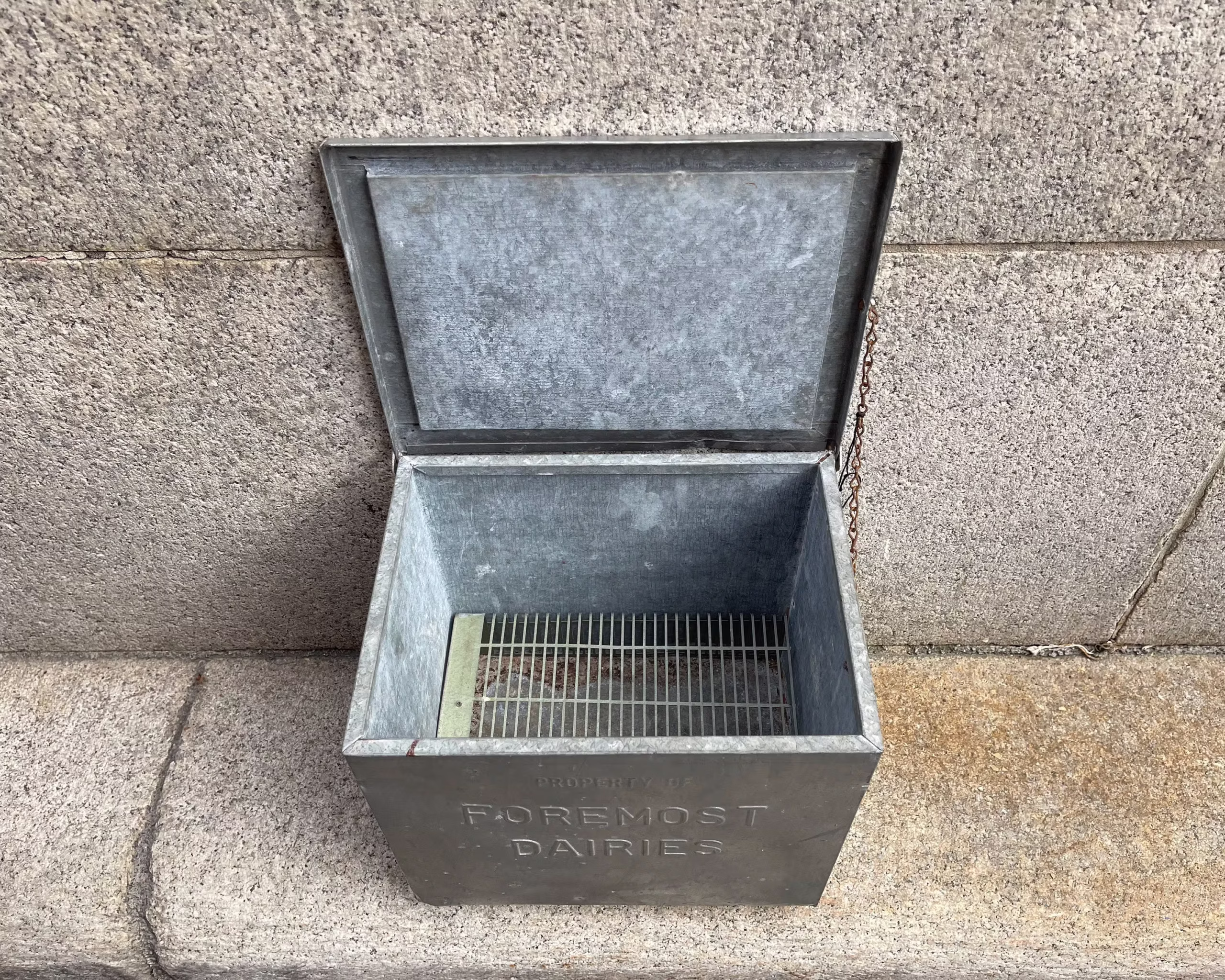 A vintage metal box with a hinged lid reading “Foremost Dairies,” sitting open on a stone surface against a stone wall. The empty box has a removable grate at the bottom.