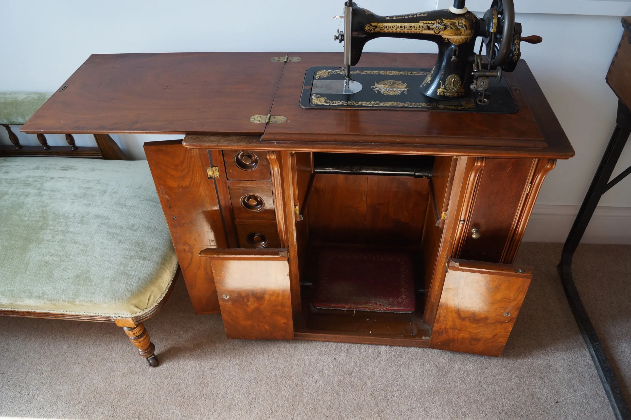 A vintage sewing machine set in a wooden cabinet with open doors, revealing storage compartments. An upholstered bench sits to the left on a beige carpeted floor.