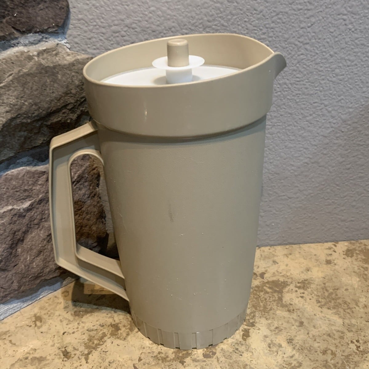 A beige plastic pitcher with a handle and white lid sits on a stone countertop next to a textured wall with stone accents.