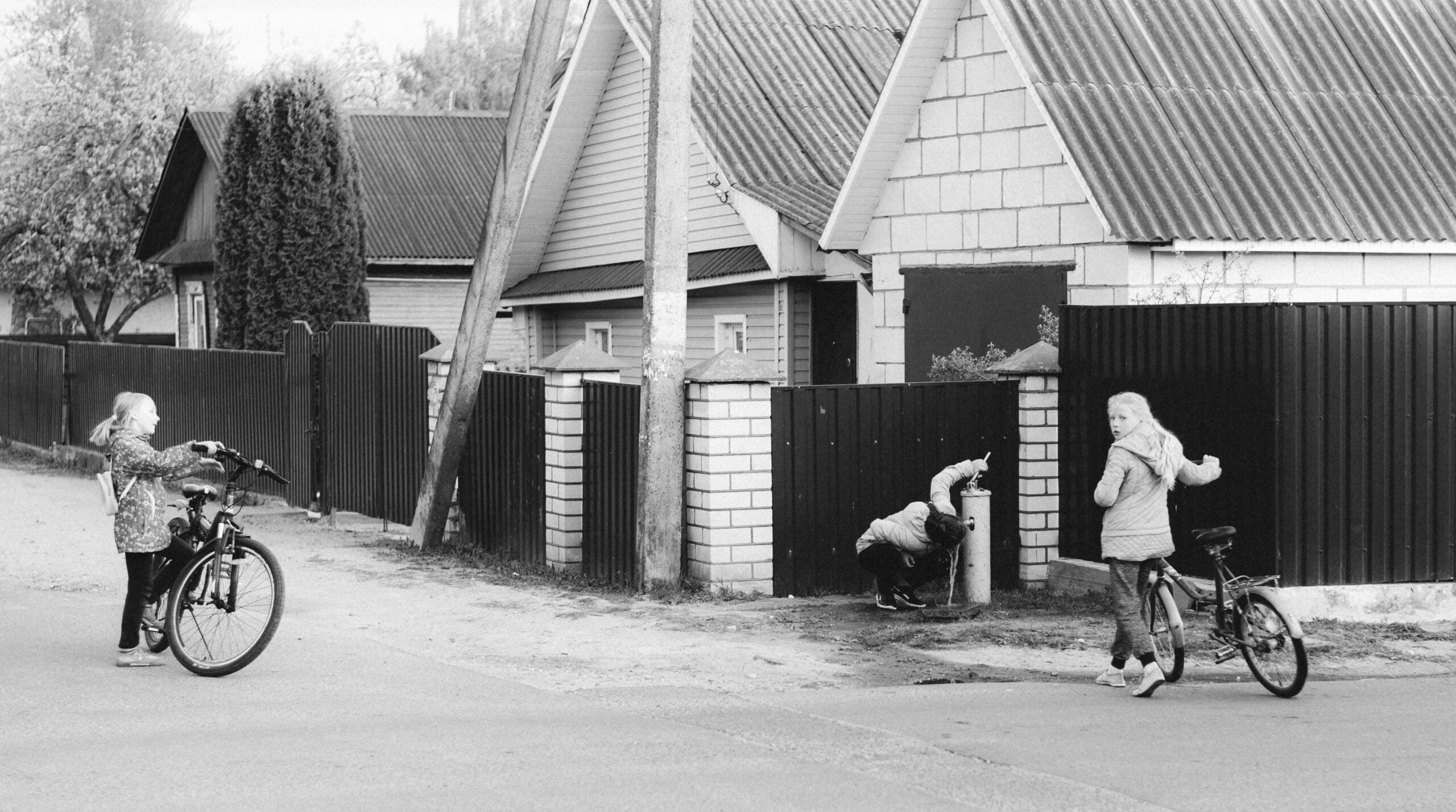 Three children with bicycles are on a quiet residential street; one drinks from a street water pump, while the other two watch. Houses with fences and trees line the background. The image is in black and white.