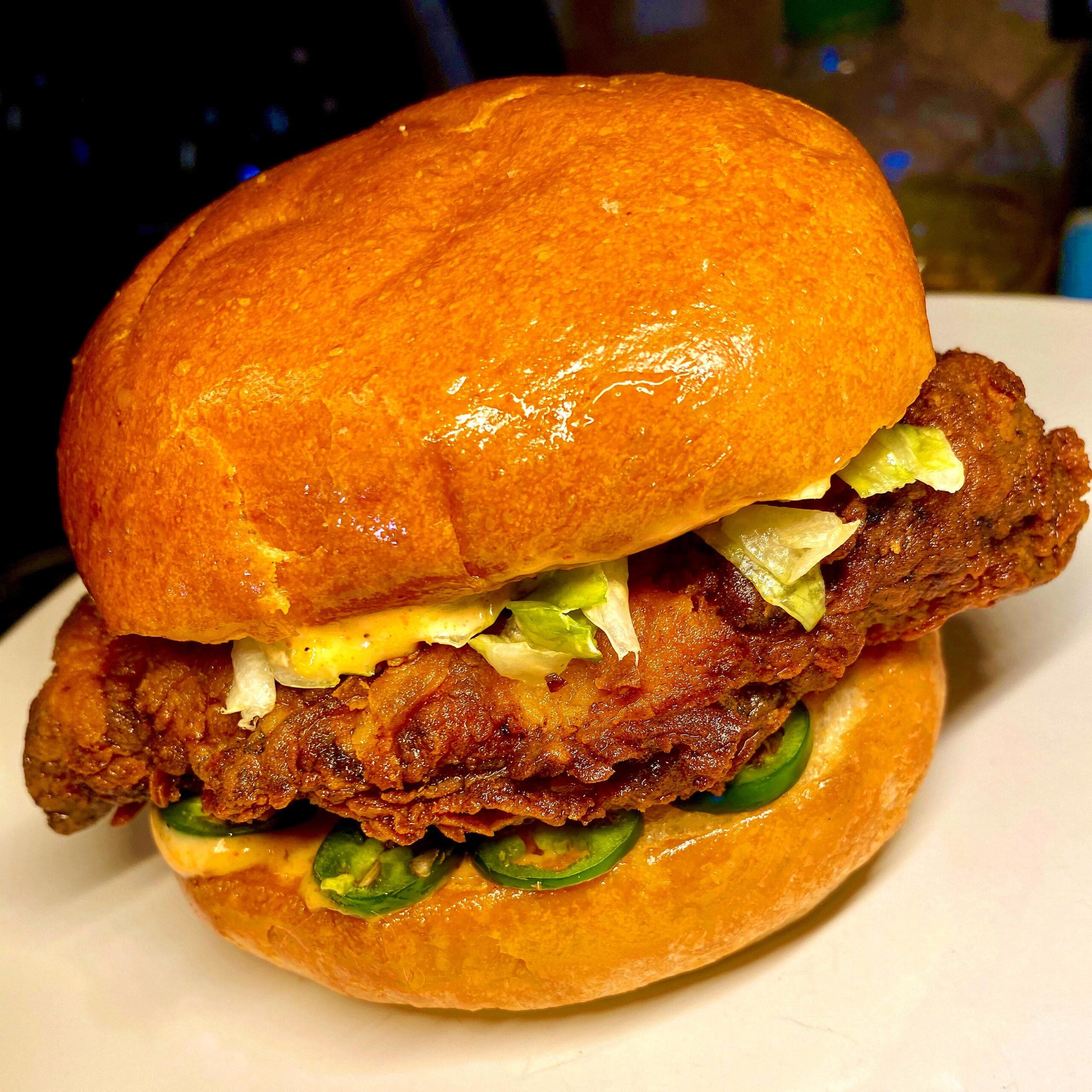 A crispy fried chicken sandwich on a golden bun with lettuce, sliced jalapeños, and creamy sauce, served on a white plate.