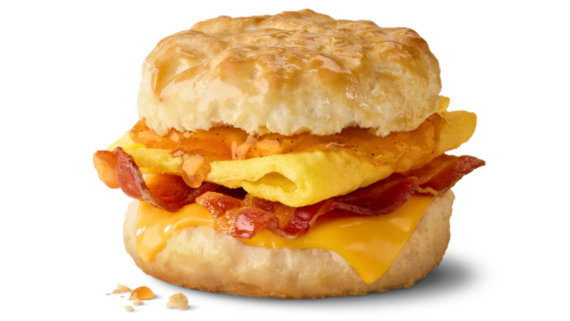 A limited-time fast-food item, this breakfast sandwich features a flaky biscuit, scrambled eggs, crispy bacon, cheddar cheese, and a golden top, shown on a white background with a few biscuit crumbs nearby.