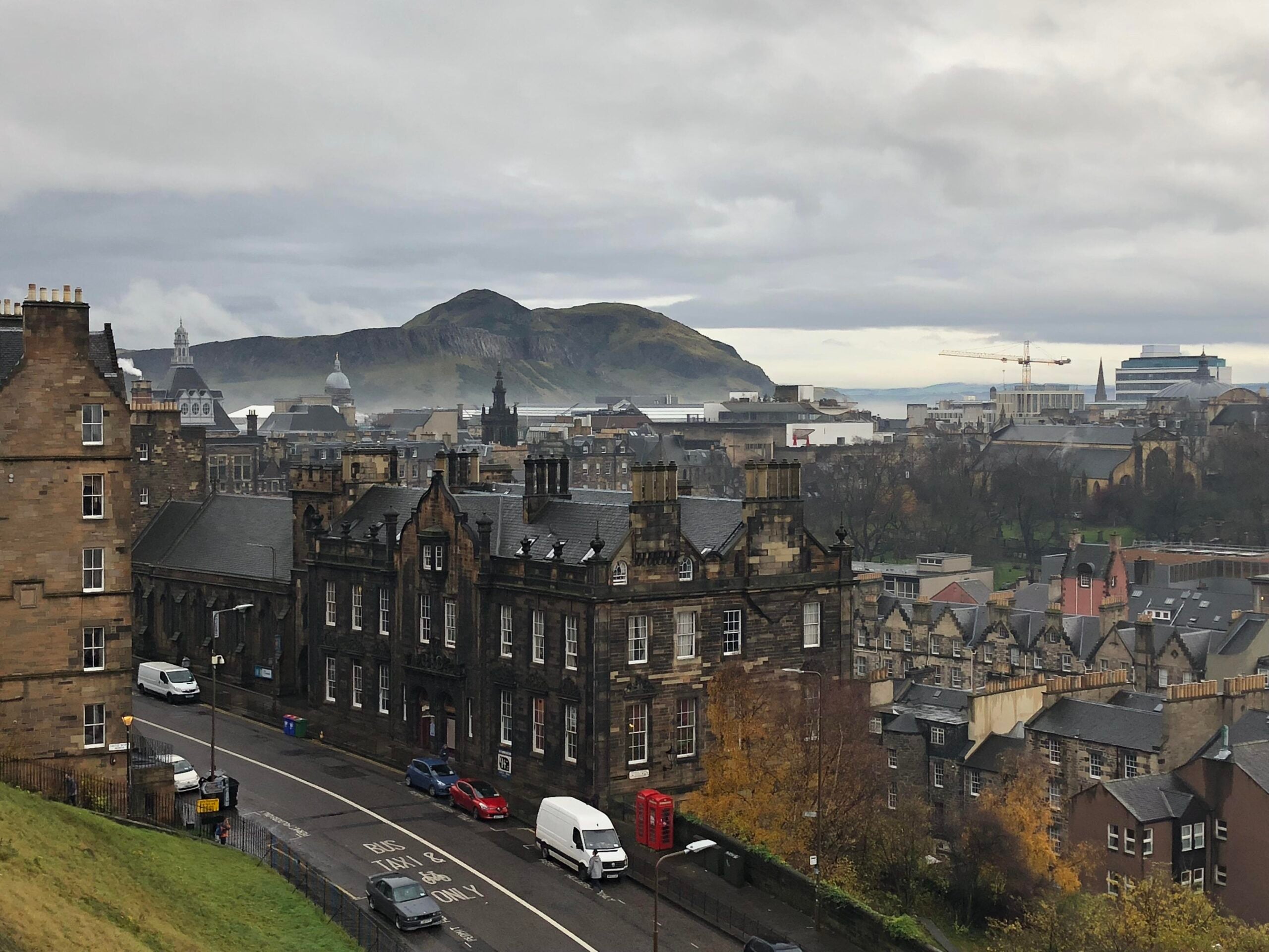 A cloudy view of historic stone buildings in Edinburgh, with Arthur’s Seat in the background. Cars are parked along the street, and a red phone booth stands near autumn trees. Cranes and a ship are visible farther back.