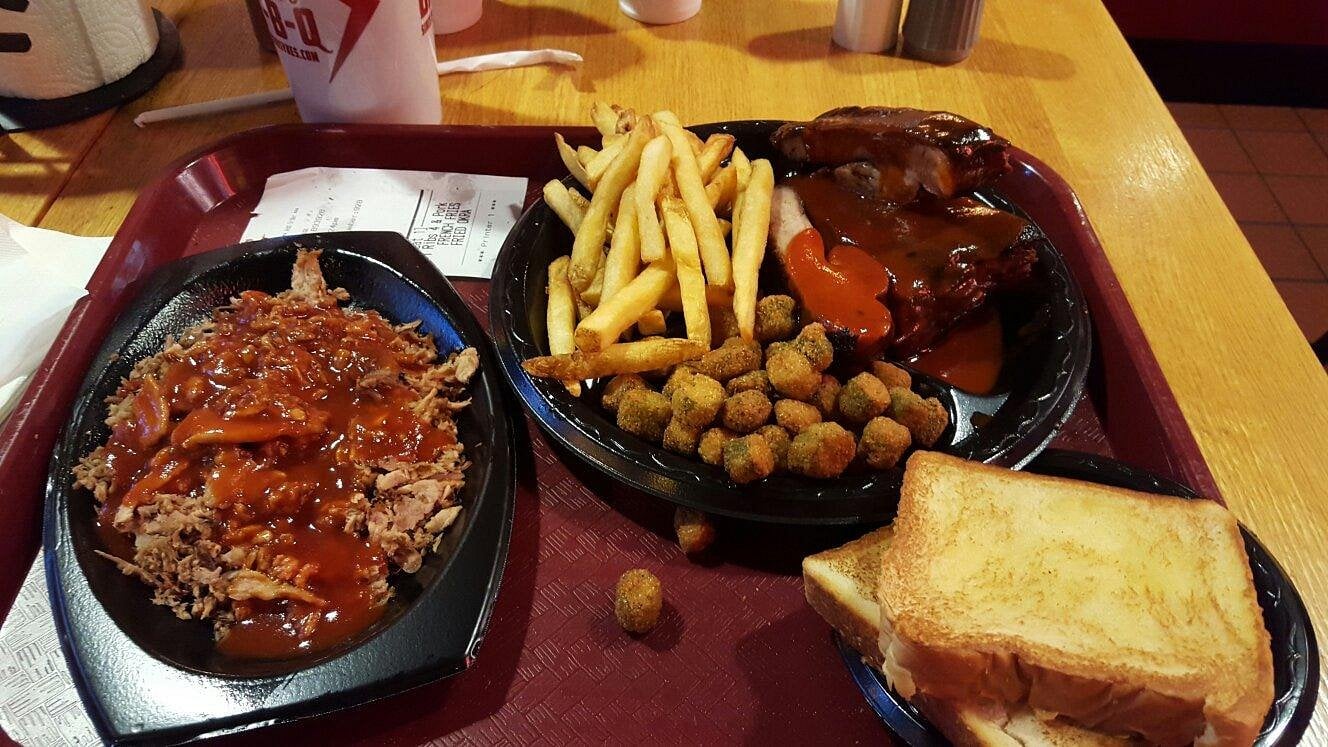 Barbecue meal with pulled pork, fries, ribs with barbecue sauce, fried okra, two slices of toasted bread, and a drink cup on a red tray on a wooden table.