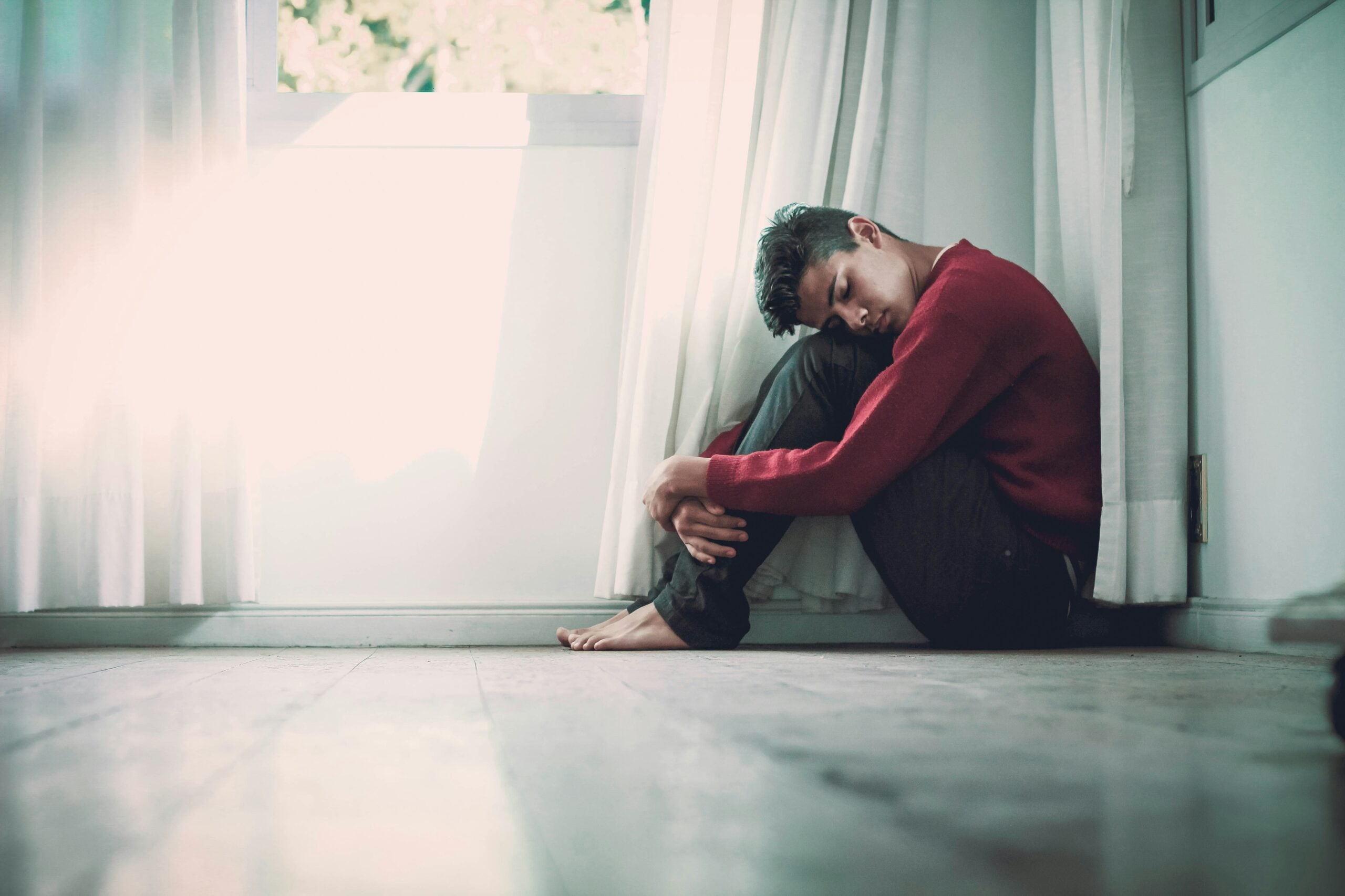 A person sits on the floor by a window, hugging their knees and looking down, appearing sad or deep in thought. White curtains and soft light fill the room, capturing a quiet moment of reflection on life in 2026.