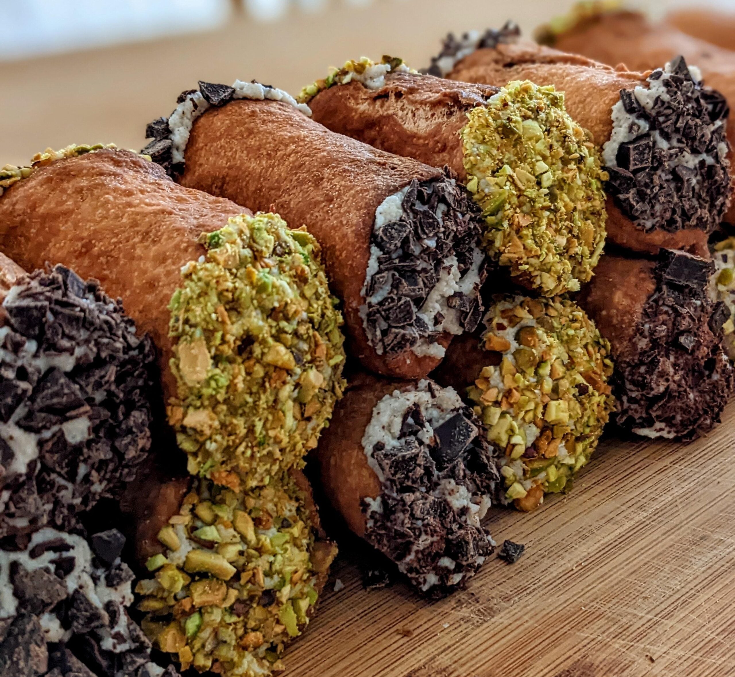 A close-up of several homemade cannolis filled with cream, with their ends dipped in chopped pistachios and chocolate, arranged on a wooden surface.