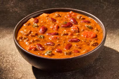 A dark bowl filled with chili, featuring red kidney beans, ground meat, tomatoes, and green peppers in a thick, reddish-brown sauce.