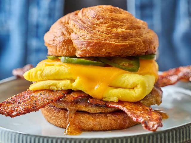 A croissant sandwich filled with folded eggs, cheddar cheese, jalapeño slices, bacon, and sausage, served on a plate.