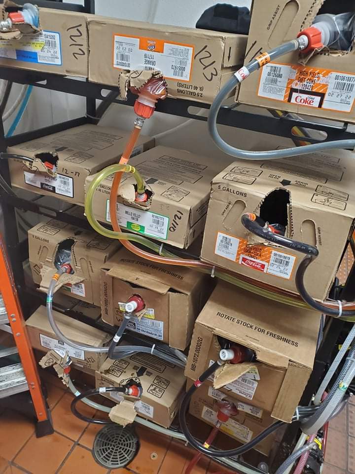 Several boxes of soda syrup are connected to hoses on a metal rack in a restaurant storage area, with labels for various soft drink brands visible on the boxes.