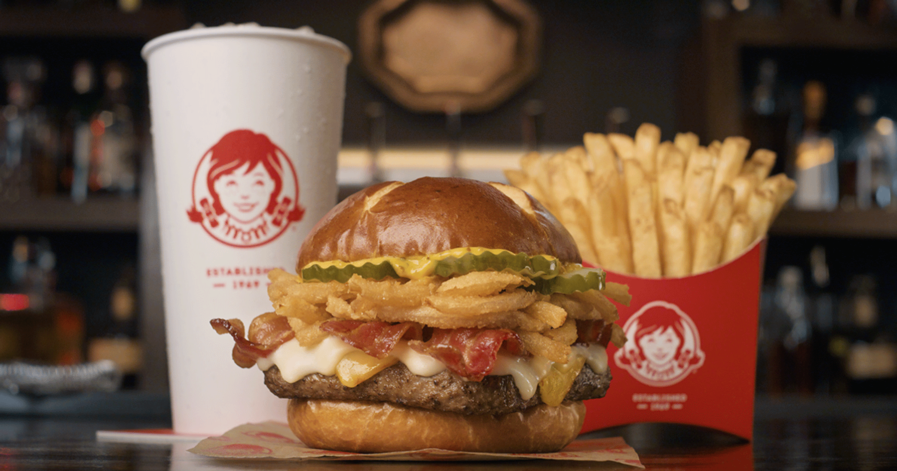 A burger with bacon, cheese, pickles, and crispy onions on a bun sits in front of a large cup and a container of fries, all in Wendy’s branded packaging on a counter. Limited-time fast-food items.