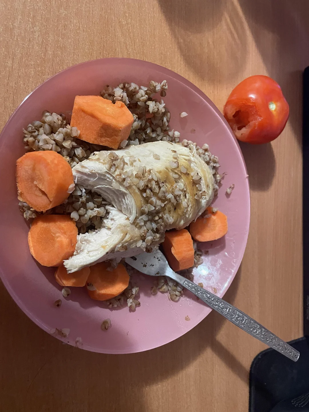A pink plate with cooked buckwheat, sliced carrots, and a piece of chicken breast. A fork rests on the plate. A partially eaten tomato is on the table beside the plate.