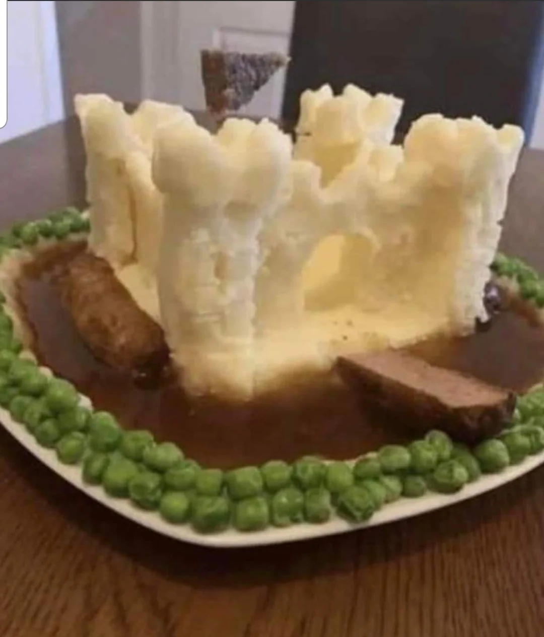 A plate of food arranged as a castle made from mashed potatoes, with meat and gravy as moat and walls, and green peas lining the edge.
