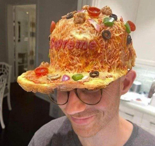 A man wearing glasses smiles while wearing a hat made to look like a pizza, with toppings and the word “supreme” spelled out in pepperoni on the front.