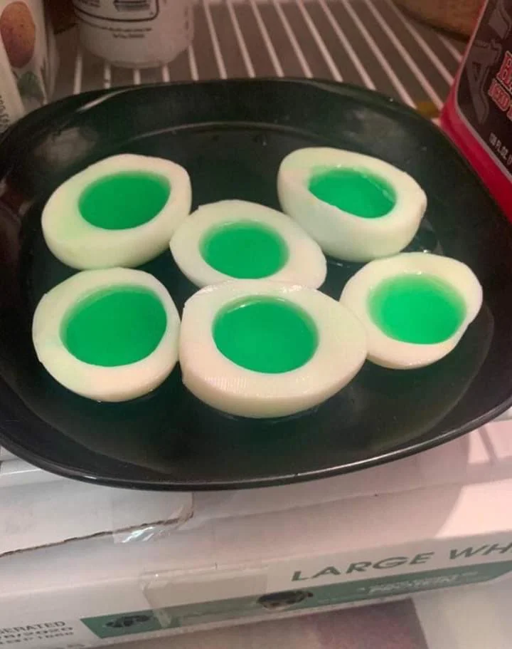 Six halved white eggs with bright green centers are arranged on a black plate inside a refrigerator, giving them an unusual, artificial appearance.