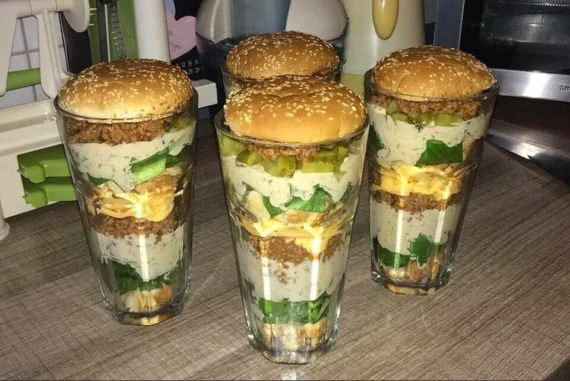 Four tall glasses are layered with burger ingredients&mdash;ground beef, cheese, pickles, lettuce, and sauce&mdash;each topped with a sesame seed bun, resembling deconstructed cheeseburgers.