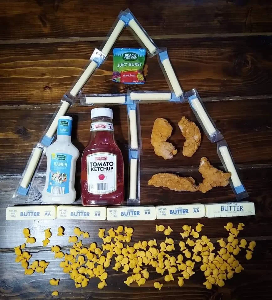 Snacks and condiments arranged in the shape of a house, with string cheese as the roof, sauces as windows, chicken tenders as people, and goldfish crackers as the ground.