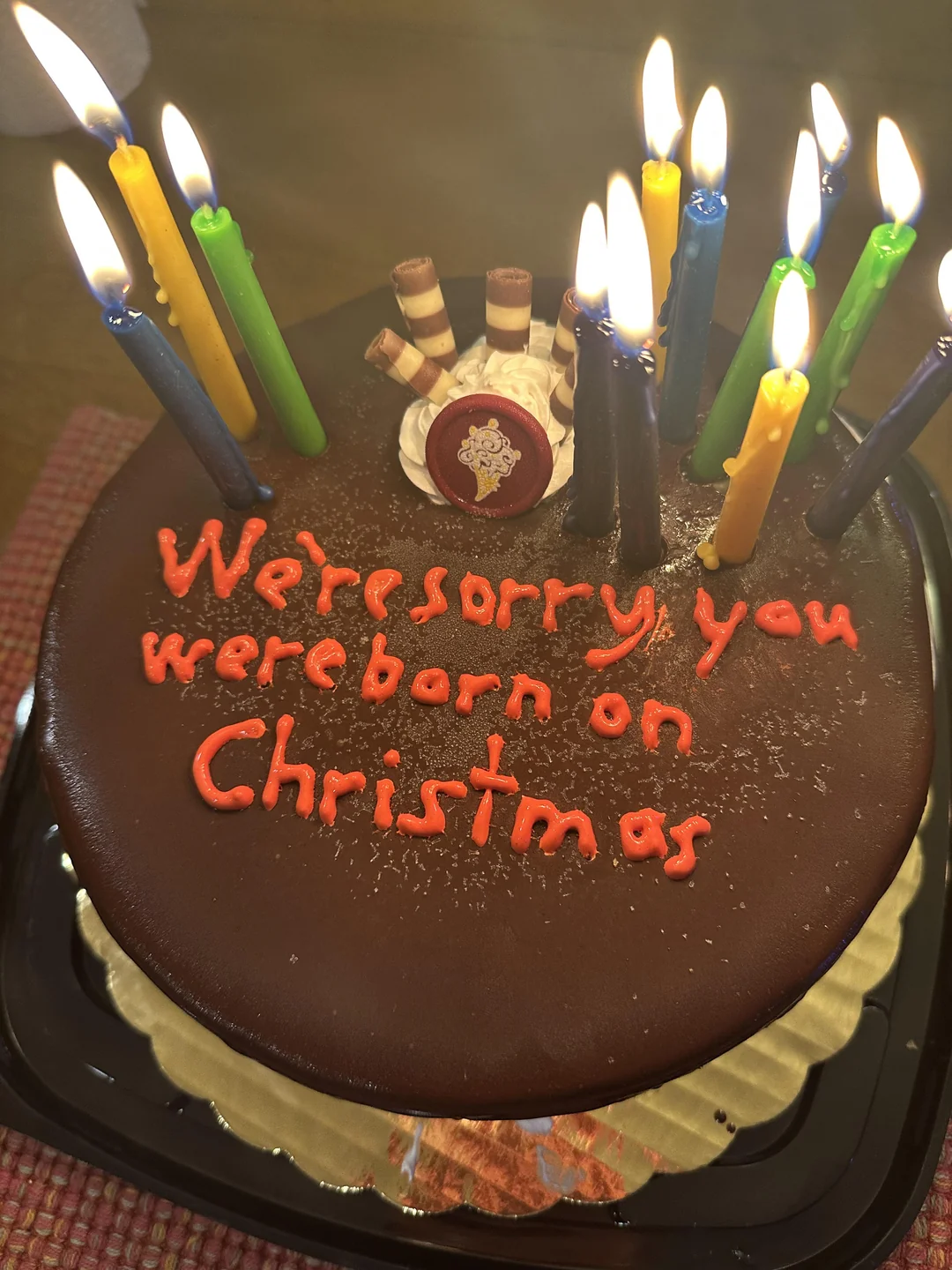 A chocolate cake with candles and red icing that says, “We’re sorry you were born on Christmas.” There are cookie sticks and a small decoration on top.