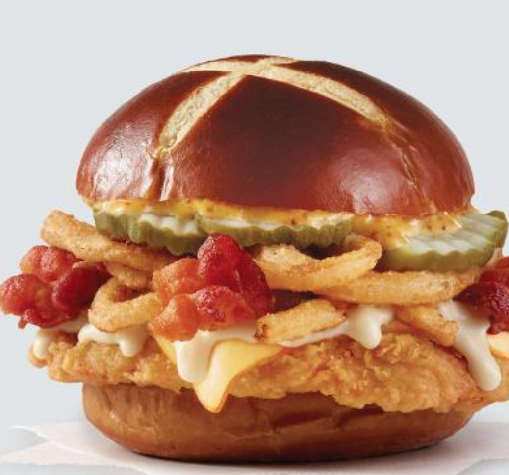A fried chicken sandwich with cheese, crispy onions, bacon, pickles, and sauce on a pretzel bun.