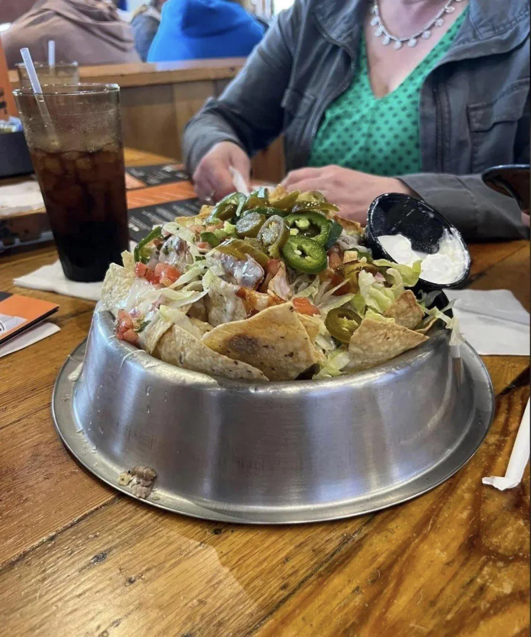 A salad with chips, lettuce, tomatoes, jalapeños, and sour cream is served in a metal dog bowl on a wooden table at a restaurant, with a drink and a person in a green dress visible.