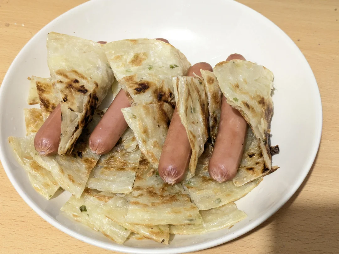 A plate with cooked hot dogs wrapped in pieces of crispy flatbread or scallion pancakes, arranged on a white dish on a light wooden surface.