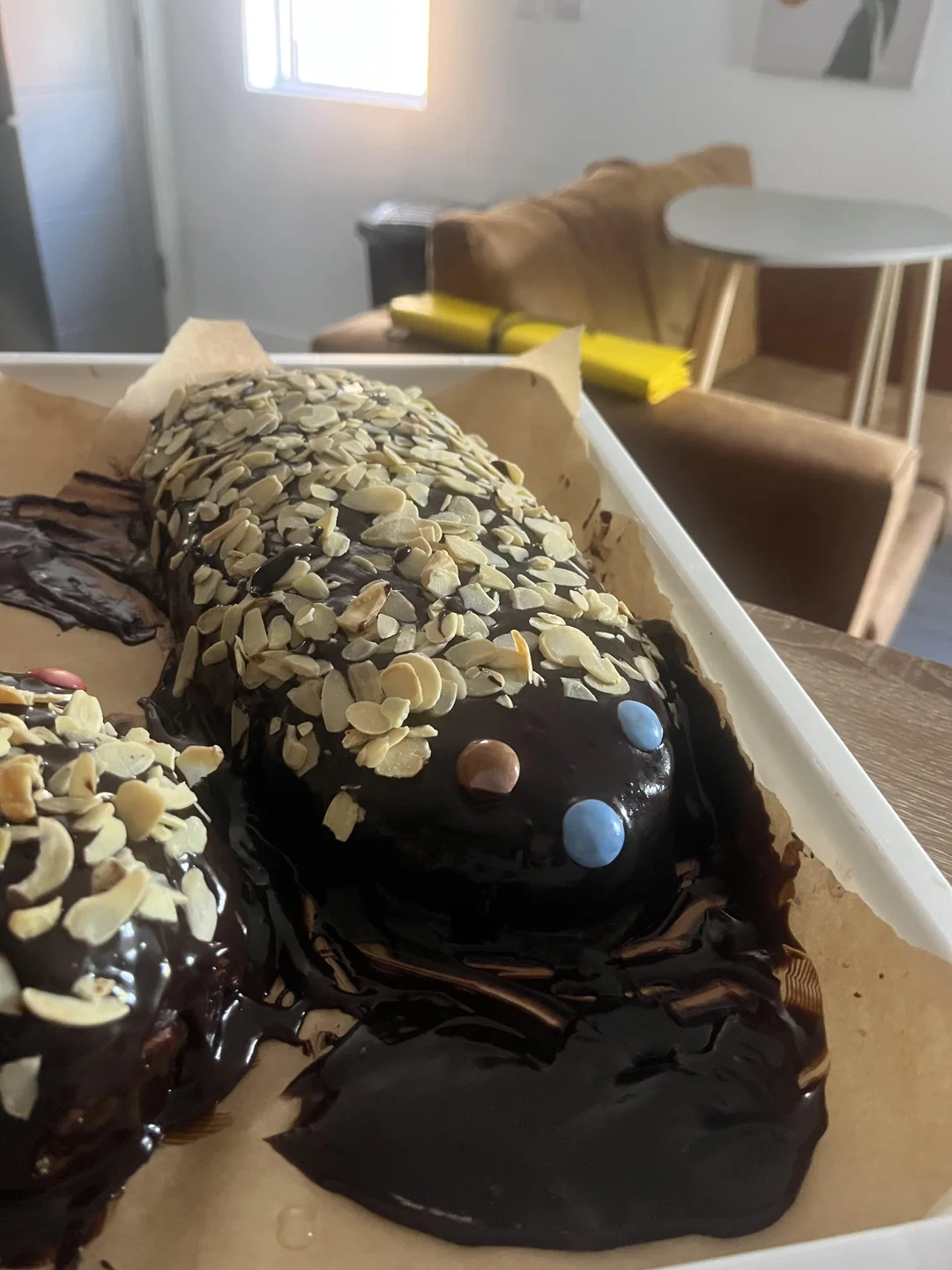 A chocolate cake shaped like a salamander, covered with sliced almonds and decorated with blue and brown candy eyes, sits on parchment paper in a home kitchen.